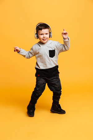 Picture of little funny boy child standing isolated over yellow background. Looking camera listening music with headphones.の写真素材