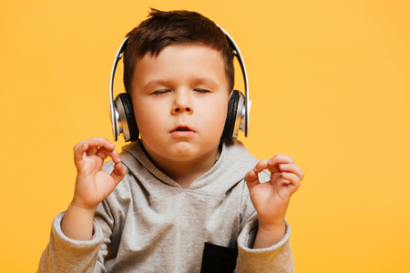 Photo of little concentrated boy child sitting on floor isolated over yellow background with eyes closed listening music with headphones.の写真素材
