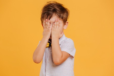 Image of little boy child standing isolated over yellow background covering face with hands.の写真素材
