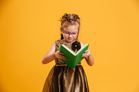 Image of concentrated girl child wearing princess crown standing isolated over yellow background. Looking aside reading book.の写真素材
