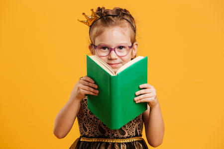 Photo of cute girl child wearing princess crown standing isolated over yellow background. Looking camera reading book.の写真素材