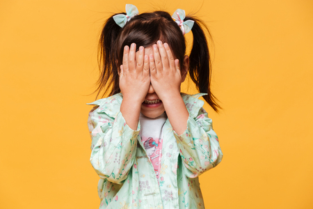 Photo of little girl child standing isolated over yellow background covering eyes with hands.の写真素材