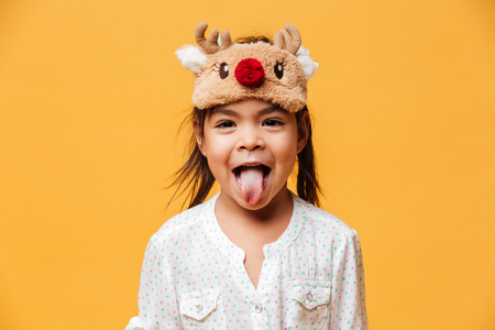 Photo of smiling little girl child standing isolated over yellow background wearing christmas cute mask showing tongue.の写真素材