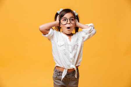 Image of shocked little girl child wearing glasses standing isolated over yellow background. Looking...