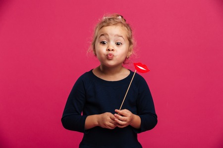 Pretty young blonde girl sends air kiss and holding paper lips while looking aside over pink backgroundの写真素材