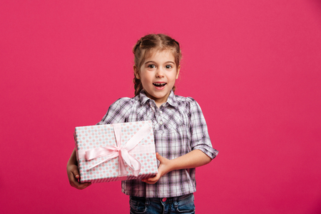 Picture of happy little girl child standing isolated over pink background holding gift box surprise.の写真素材