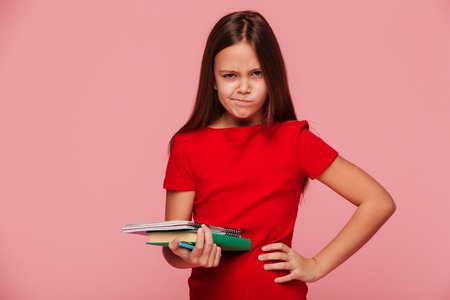 Portrait of displeased girl in red dress holding book and looking camera isolated over pinkの写真素材