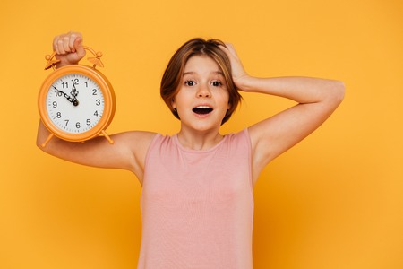 Surprised smiling girl in pink dress showing alarm clock and holding her head isolated over yellowの写真素材