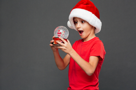 Photo of surprised little boy child wearing christmas santa hat standing isolated over grey background. Looking aside holding christmas toy.の写真素材