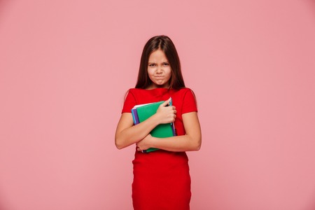 Unhappy displeased girl in red dress holding book and looking camera isolated over pinkの写真素材
