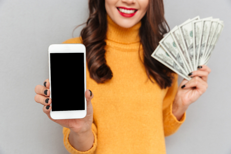Cropped image of smiling brunette woman in sweater holding money and showing blank smartphone screen over gray backgroundの写真素材