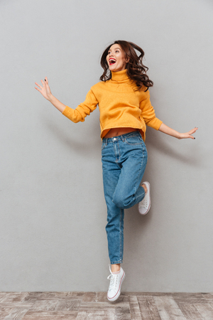 Full length image of Happy brunette woman in sweater jumping and looking away over gray backgroundの写真素材
