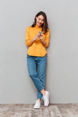 Full length image of Happy brunette woman in sweater writing message on smartphone over gray backgroundの写真素材