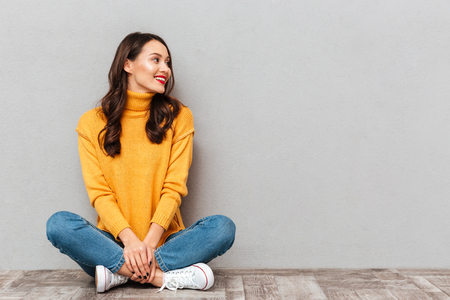 Happy brunette woman in sweater sitting on the floor and looking away over gray backgroundの写真素材
