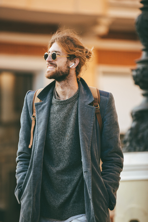 Portrait of a happy bearded man wearing sunglasses and coat walking on a city street and looking awayの写真素材