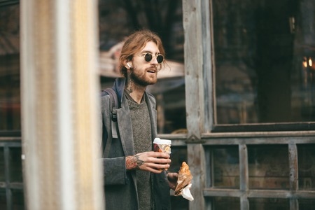 Portrait of a young bearded man dressed in coat holding croissant and cup of coffee while walking outdoors on a city streetの写真素材