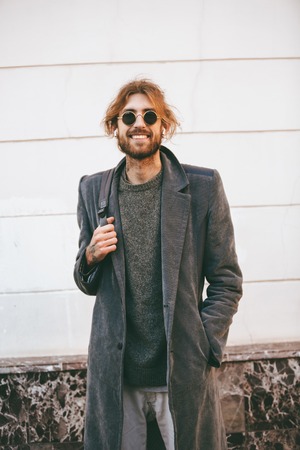 Portrait of a happy bearded man wearing sunglasses and coat standing on a city street outdoorsの写真素材
