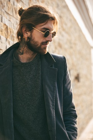 Portrait of a confident bearded man wearing sunglasses and coat leaning on a wall outdoors