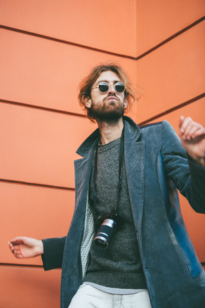 Portrait of a trendy bearded man with camera wearing sunglasses and coat standing against a wall on a city streetの写真素材