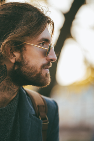 Close up portrait of a handsome bearded man with backpack wearing sunglasses and coat walking on a city streetの写真素材