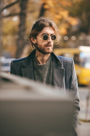 Portrait of a handsome bearded man in sunglasses and coat walking on a city streetの写真素材