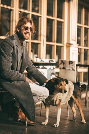 Portrait of a happy bearded man dressed in coat sitting outdoors and petting a dogの写真素材