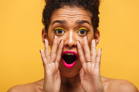 Closeup photo of african american woman emotionally shouting or calling on camera putting hands at mouth isolated over yellow wallの写真素材