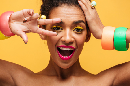 Closeup portrait of playful mulatto woman with yellow eyelids and pink lips gesturing two fingers at...
