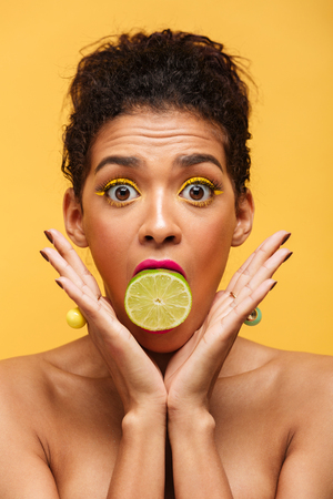 Vertical image of surprised afro american woman bulging eyes while putting half of fresh lime in mouth isolated over yellow backgroundの写真素材