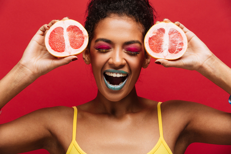 Food fashion photo of delighted afro american woman having fun holding two halves of fresh ripe grapefruit at face isolated over red wallの写真素材