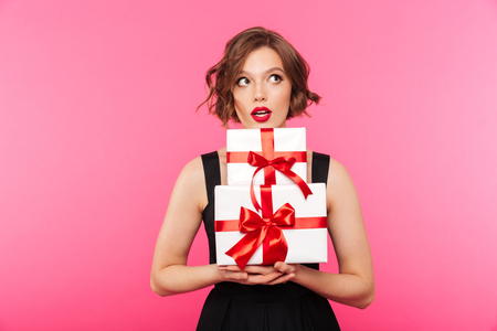 Portrait of a pretty girl dressed in dress holding stack of gift boxes and looking away isolated over pink backgroundの写真素材