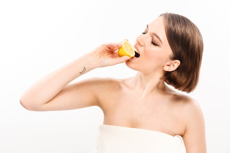 Beauty portrait of a young woman with short brunette hair wrapped in towel eating sliced lemon isolated over white backgroundの写真素材