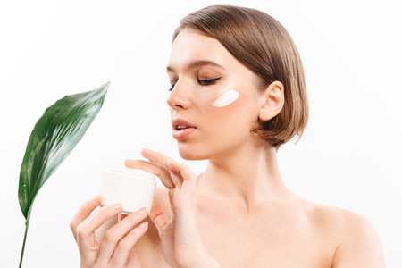 Beauty portrait of a sensual half naked woman with short brunette hair posing with green leaves and holding cream container isolated over white backgroundの写真素材