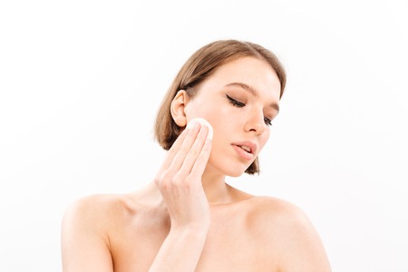 Beauty portrait of a healthy shirtless woman with short brunette hair removing makeup with a cotton pad isolated over white backgroundの写真素材