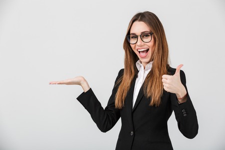 Portrait of a cheerful businesswoman dressed in suit showing thumbs up and holding copy space on her palm isolated over gray backgroundの写真素材