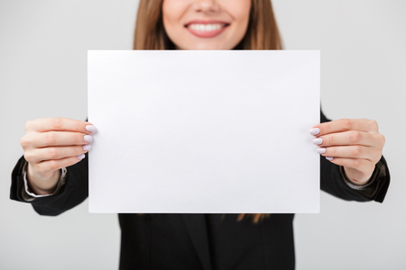 Close up of a smiling businesswoman dressed in suit showing blank piece of paper isolated over gray backgroundの写真素材