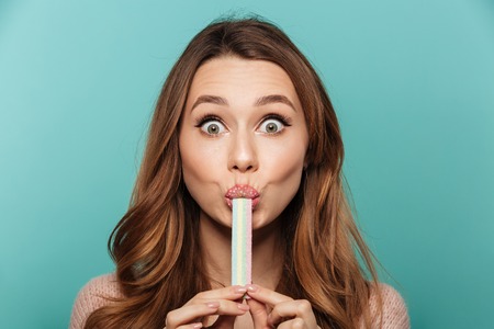 Beauty portrait of a cute brown haired woman with sparkling makeup eating sugar candy isolated over blue backgroundの写真素材