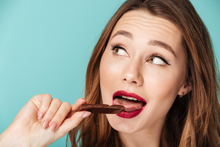 Portrait of a satisfied brown haired woman with bright makeup eating chocolate bar isolated over blue backgroundの写真素材