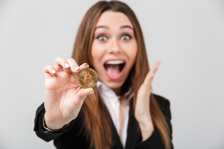 Happy amazed businesslady with opened mouth looking camera and holding golden bitcoin isolated over grey wallの写真素材