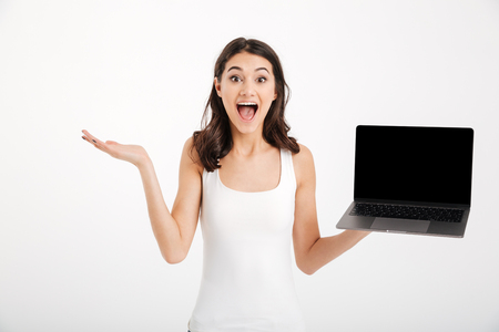 Portrait of an excited girl dressed in tank-top showing blank screen laptop computer while standing and looking at camera isolated over white backgroundの写真素材