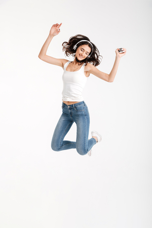 Full length portrait of a cheerful girl dressed in tank-top listening to music with headphones while holding mobile phone and jumping isolated over white backgroundの写真素材