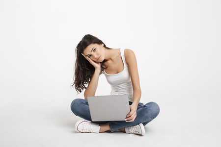 Portrait of an upset girl dressed in tank-top holding laptop while sitting on the floor isolated over white backgroundの写真素材
