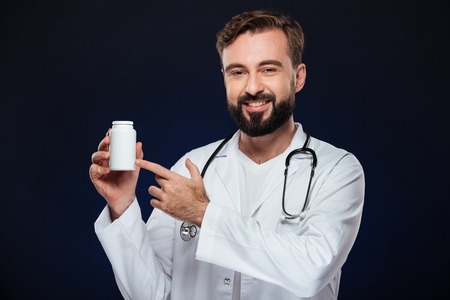 Portrait of a confident male doctor dressed in uniform with stethoscope pointing finger at bottle with pills isolated over dark backgroundの写真素材