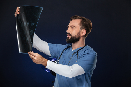 Portrait of a concentrated male doctor dressed in uniform with stethoscope examining x-ray picture isolated over dark backgroundの写真素材