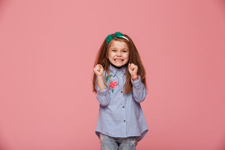 Smiling girl model in hair hoop and fashion clothes expressing happiness, gesturing with clenched fists against pink backgroundの写真素材