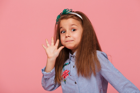 Portrait of funny female kid having long auburn hair looking on camera giving high five, meaning hi or bye with hand over pink backgroundの写真素材