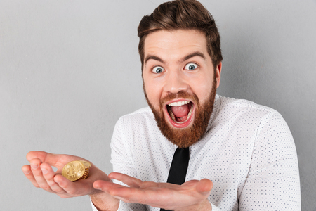 Portrait of an excited businessman showing golden bitcoins in his arms isolated over gray backgroundの写真素材