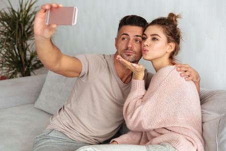 Pretty lovely couple sitting on couch and making selfie on smartphone at homeの写真素材