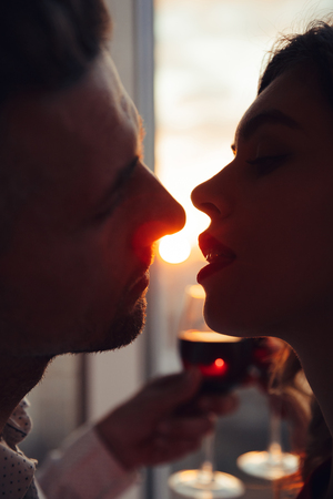 Portrait of young beautiful lovers kissing at sunset and holding glass with wine at homeの写真素材