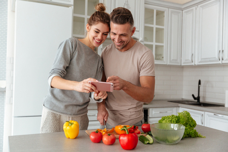 Portrait of a laughing loving couple cooking salad together while standing on a kitchen at home and pointing finger at mobile phoneの写真素材
